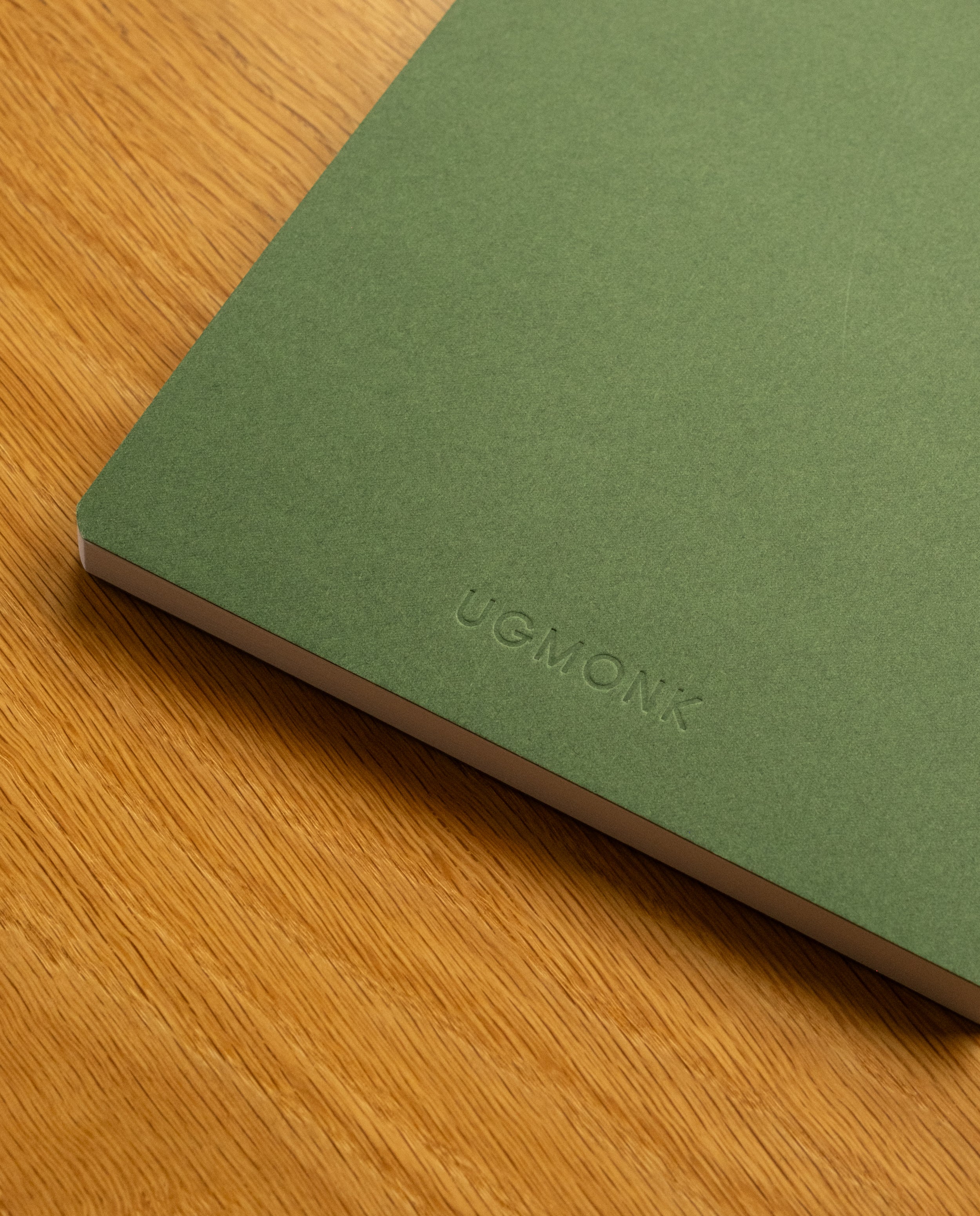 A Layflat Notebook (Green) with UGMONK embossed on the cover and Smyth sewn binding rests on a wooden surface.