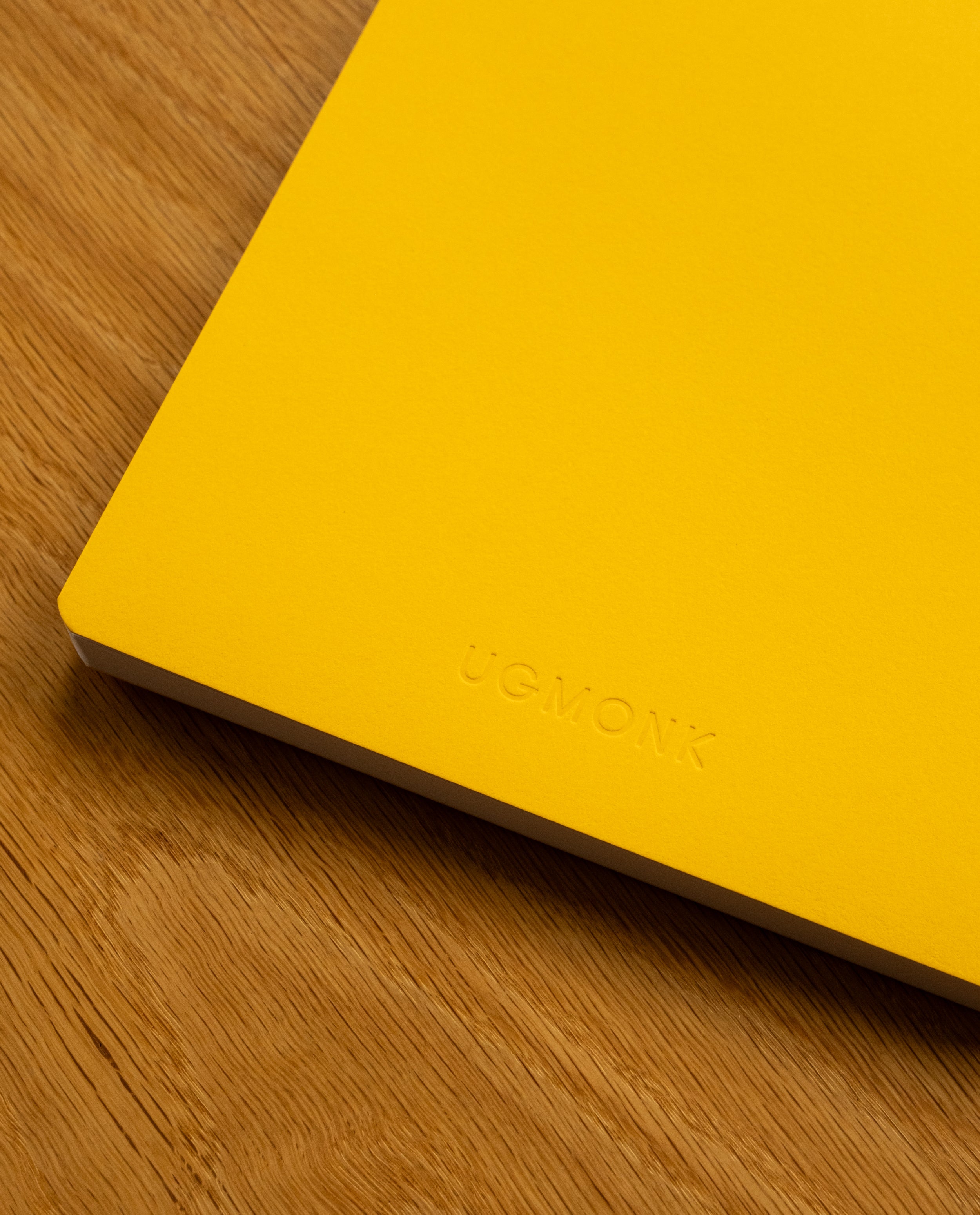 A Layflat Notebook (Yellow) with UGMONK embossed on the cover rests on a wooden surface, featuring dot grid pages and durable Smyth sewn binding.