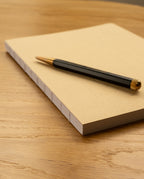 A black and gold pen rests on top of a closed Layflat Notebook (Tan) with visible Smyth sewn binding, placed on a wooden table.
