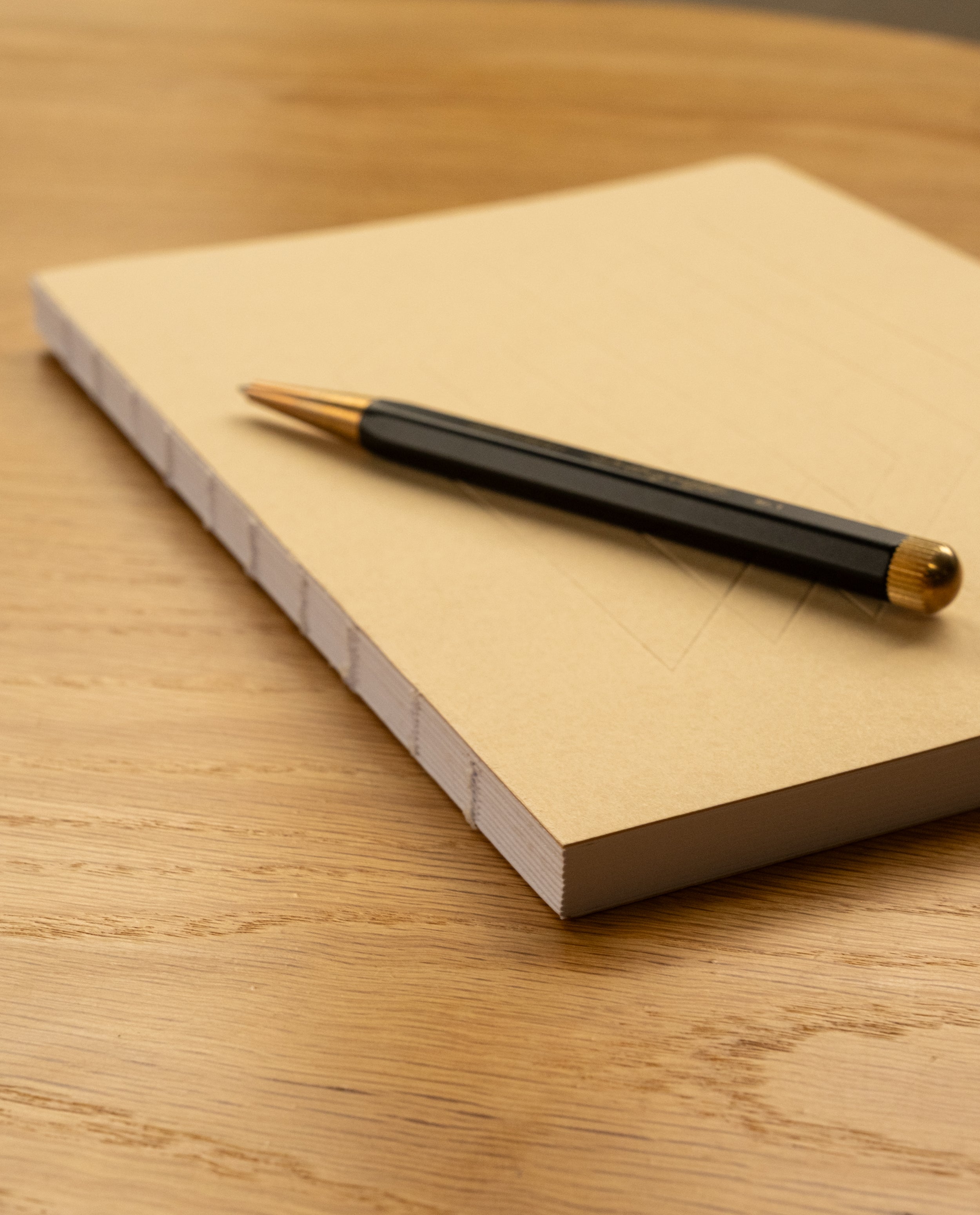A black and gold pen rests on top of a closed Layflat Notebook (Tan) with visible Smyth sewn binding, placed on a wooden table.