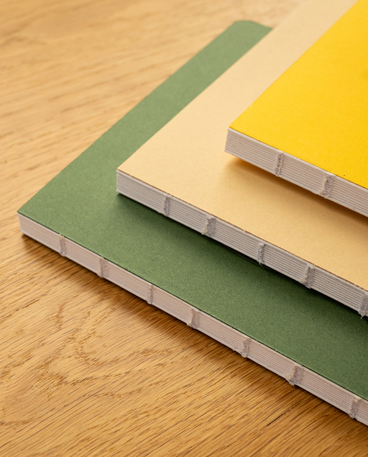 Three Layflat Notebooks (Yellow) with exposed Smyth sewn bindings and colored covers—green, beige, and yellow—are stacked on a wooden surface.