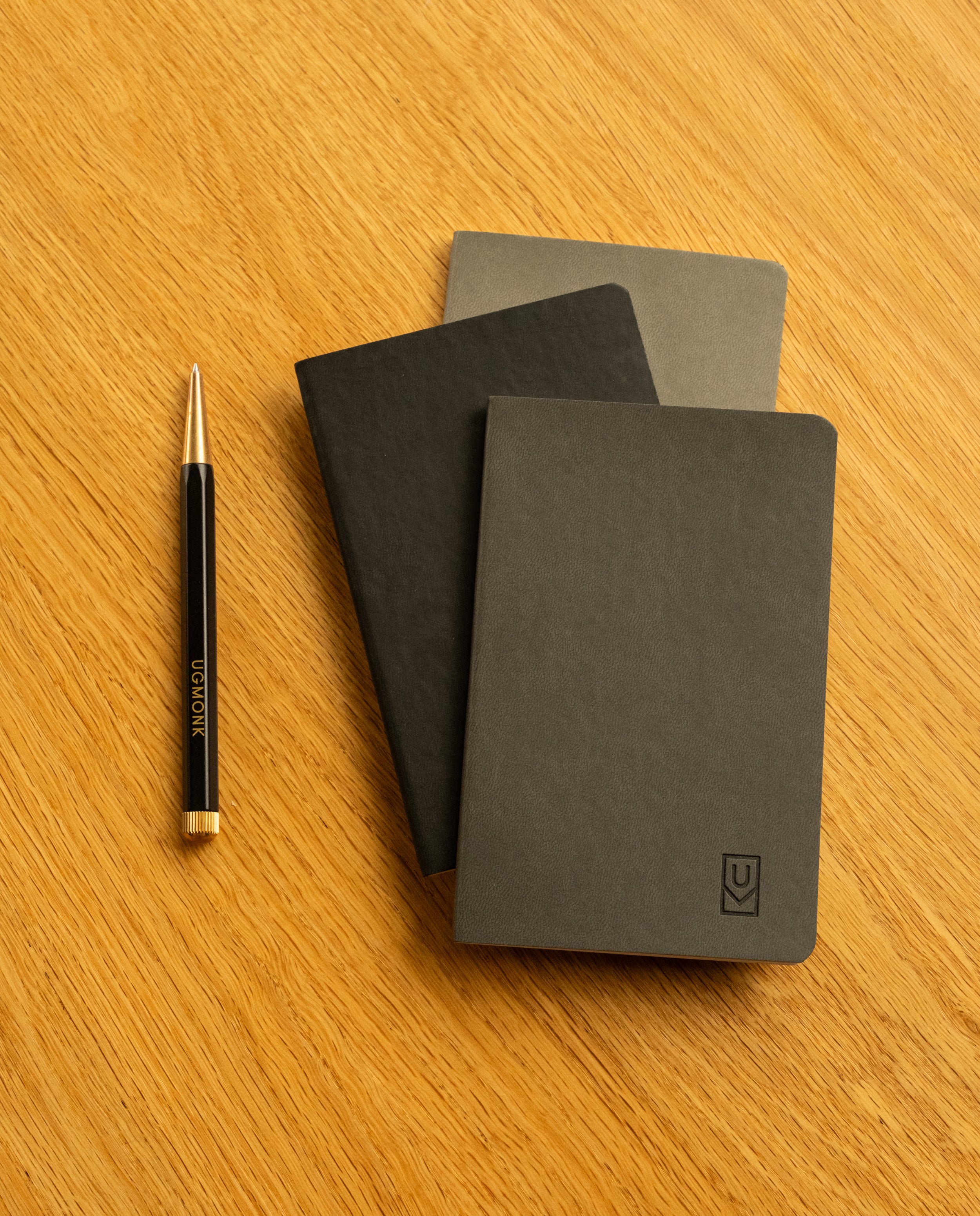A set of three Ugmonk Pocket Notebooks with black and gray soft touch covers is stacked on a wooden surface. To the left, a black pen with gold accents and "UJORK" printed on it lies ready for notes or sketches on the notebooks' dot grid pages.