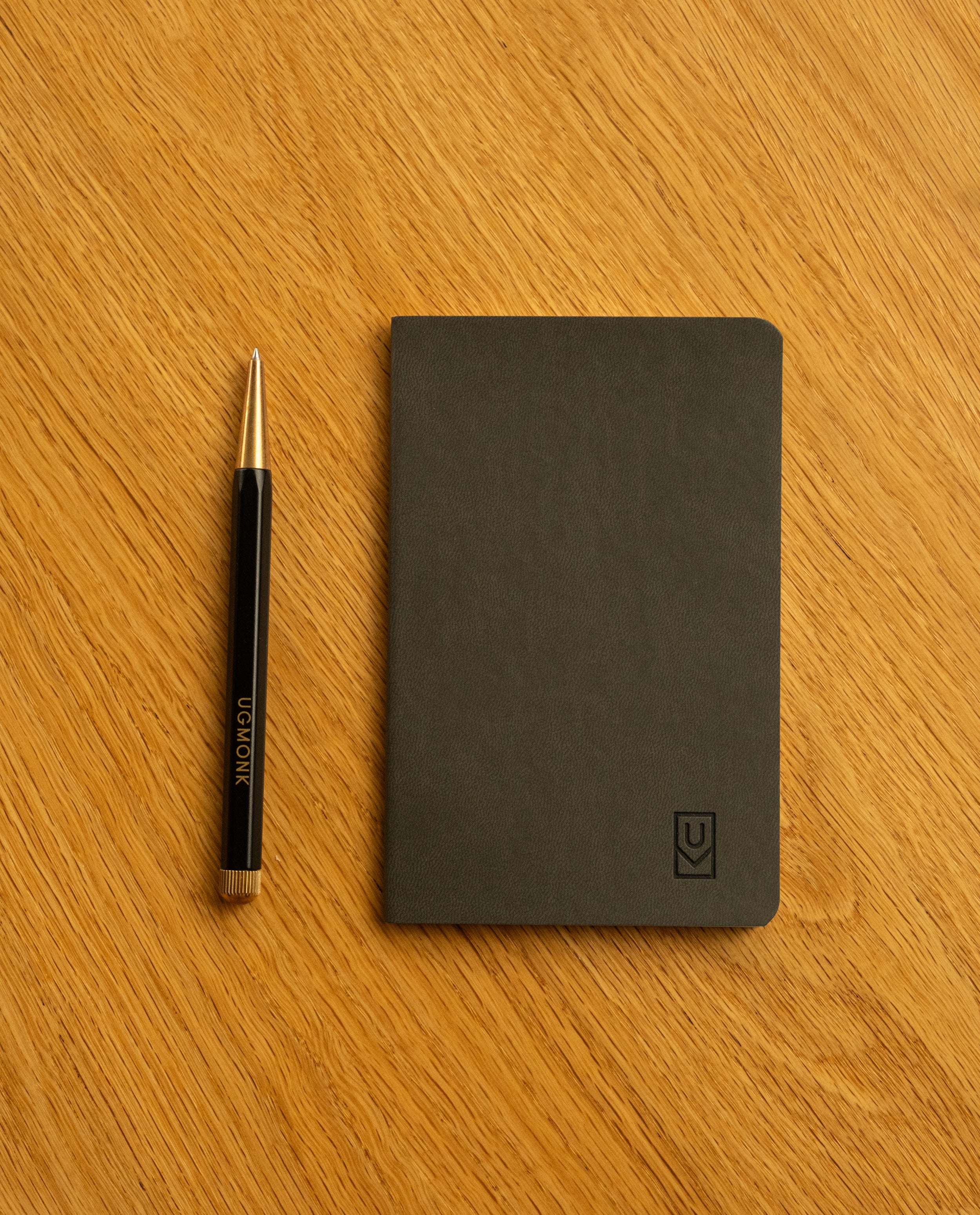 A black pen and an Ugmonk Pocket Notebook from the Set of 3 sit side by side on a light wooden surface. The small, black notebook features dot grid pages and a soft touch cover, offering both practicality and a sleek, modern design.