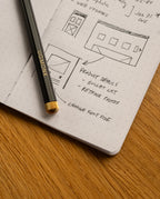 A black pencil labeled UGMONK rests on an open Ugmonk Pocket Notebook with dot grid pages, featuring sketches of webpage layouts and handwritten notes on design changes. The notebook, part of the Ugmonk Pocket Notebooks - Set of 3 with a soft touch cover, is placed on a wooden surface.