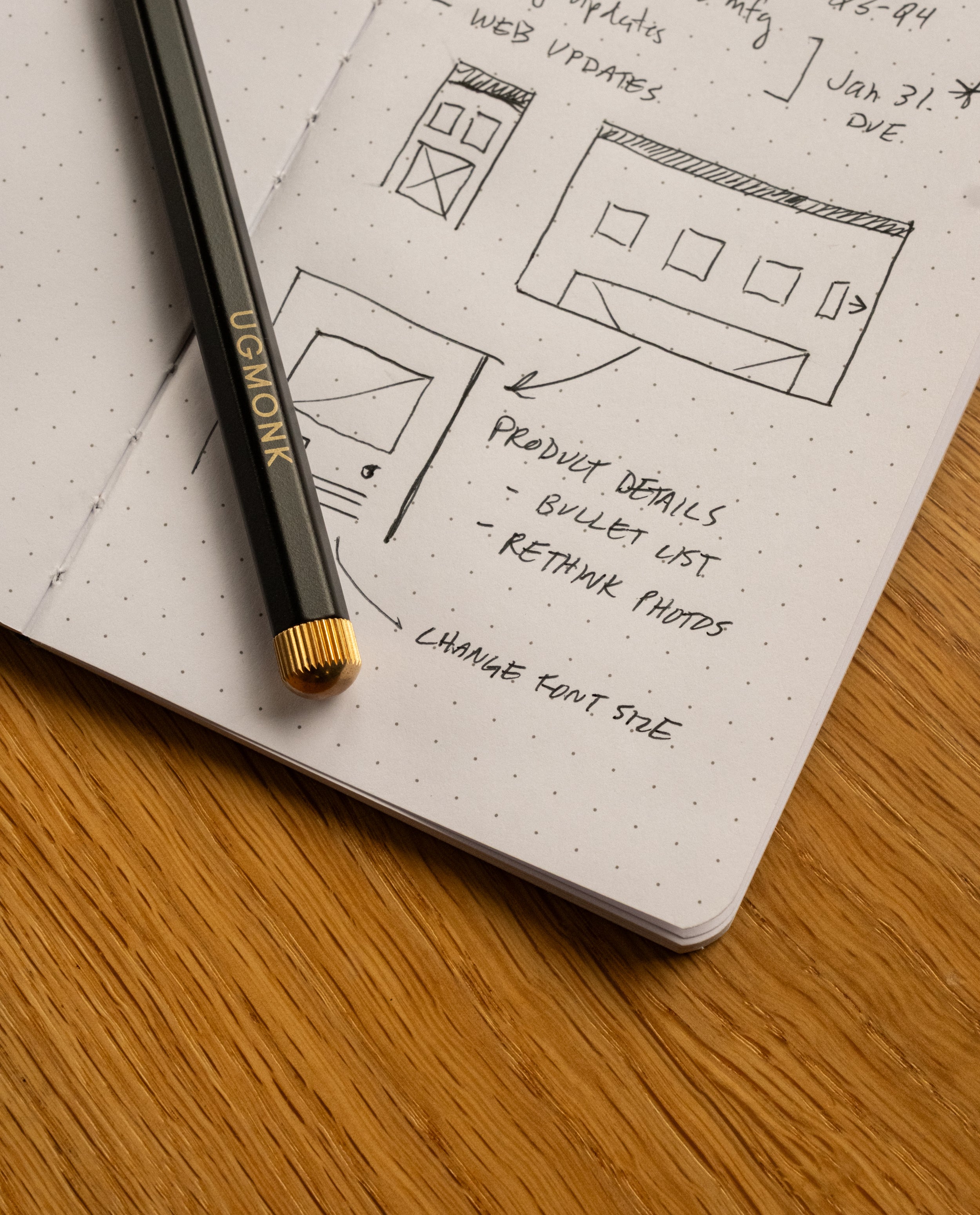 A black pencil labeled UGMONK rests on an open Ugmonk Pocket Notebook with dot grid pages, featuring sketches of webpage layouts and handwritten notes on design changes. The notebook, part of the Ugmonk Pocket Notebooks - Set of 3 with a soft touch cover, is placed on a wooden surface.