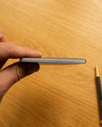 A hand holds one of the Ugmonk Pocket Notebooks - Set of 3, viewed from the side with a few pages visible. In the background, a black and gold pen rests on a wooden surface, while the notebook’s soft touch cover lends a refined feel to this everyday essential.