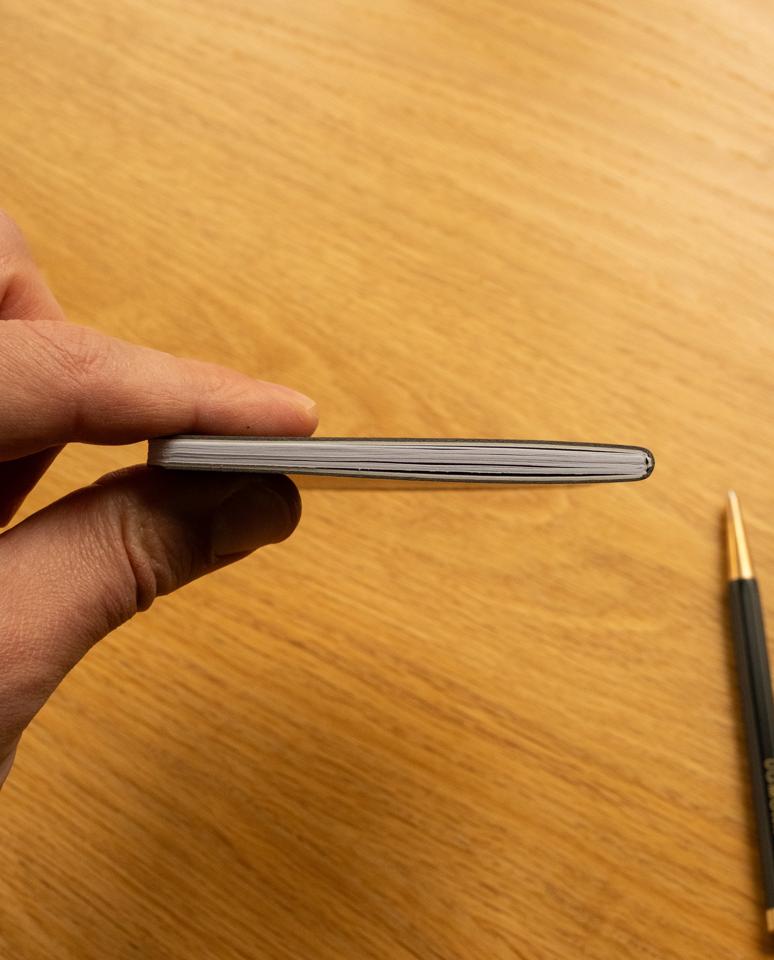 A hand holds one of the Ugmonk Pocket Notebooks - Set of 3, viewed from the side with a few pages visible. In the background, a black and gold pen rests on a wooden surface, while the notebook’s soft touch cover lends a refined feel to this everyday essential.