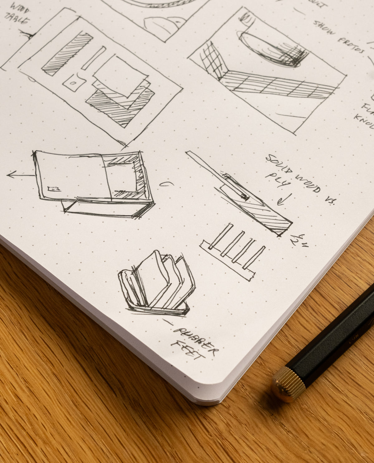 A close-up of the Layflat Notebook (Yellow) showcases pen sketches of geometric shapes and design ideas on its dot grid pages, resting on a wooden surface next to a black and gold pen. The notebook features Smyth sewn binding, ensuring it stays open for effortless creativity.
