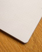 A close-up of the corner of the Layflat Notebook (Yellow) resting on a wooden surface. The page showcases its precise light gray dot grid pattern.