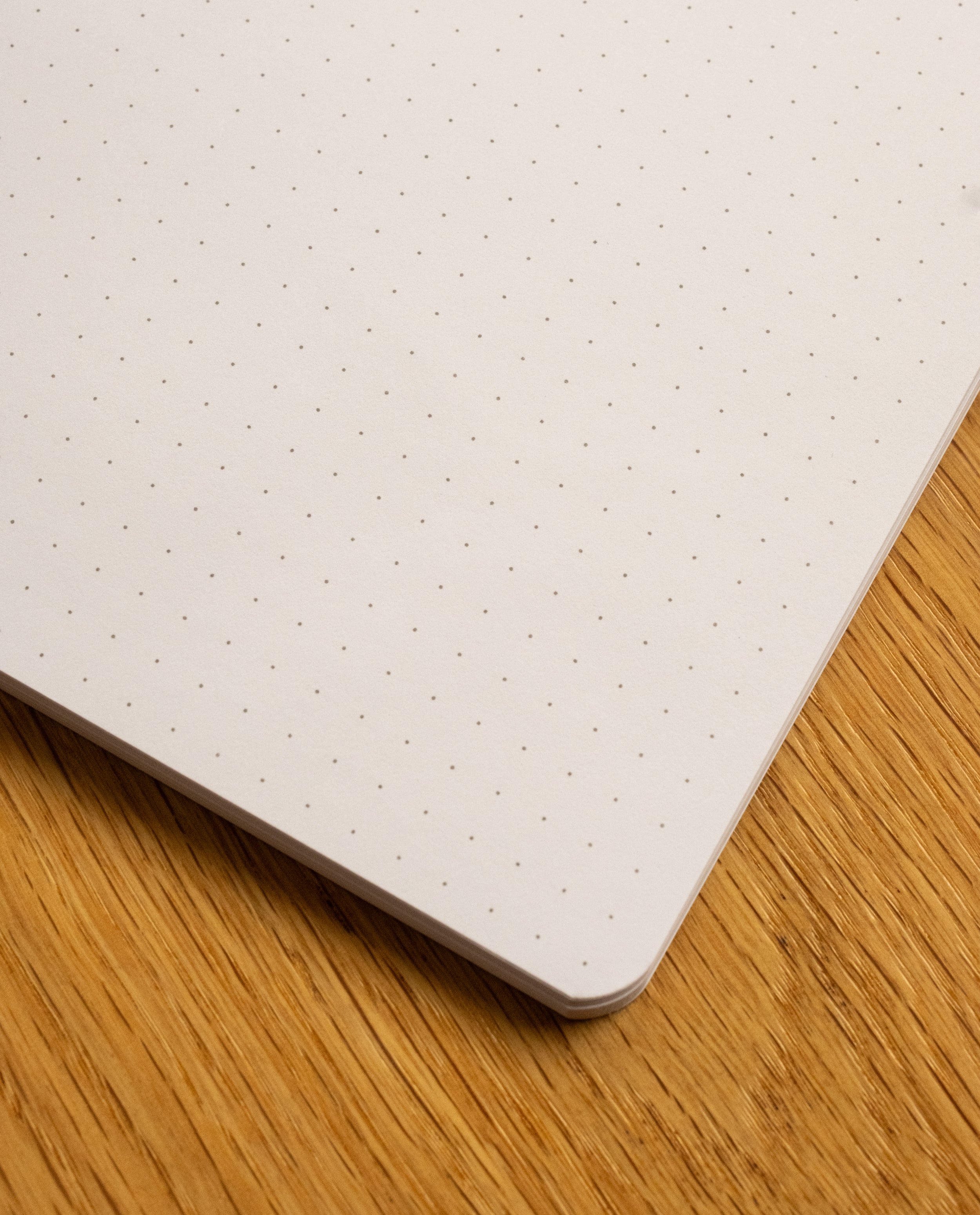 A close-up of the corner of the Layflat Notebook (Yellow) resting on a wooden surface. The page showcases its precise light gray dot grid pattern.