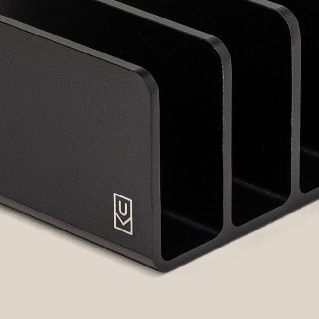 Close-up of a black metal desk organizer with three vertical slots, placed on a light beige surface. A small white logo is visible in the lower left corner of the organizer.
