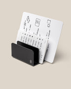 The Analog Card Organizer (Black Aluminum) holds white cards with handwritten notes, sketches, and diagrams neatly in slots. The cards feature lists, labels, and small illustrations. The background is plain and light-colored.