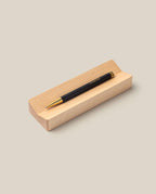 A stylish black and gold gel ink pen from The Pen & Pen Tray Bundle (Black/Maple) rests on a light maple pen tray against a beige background.