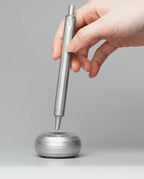 A hand holds the HMM Paperweight Pen Stand (Raw) vertically above its round, silver aluminum base on a gray background. The tip of the pen is near the center of the base, which serves as an elegant paperweight and a stylish desk accessory.