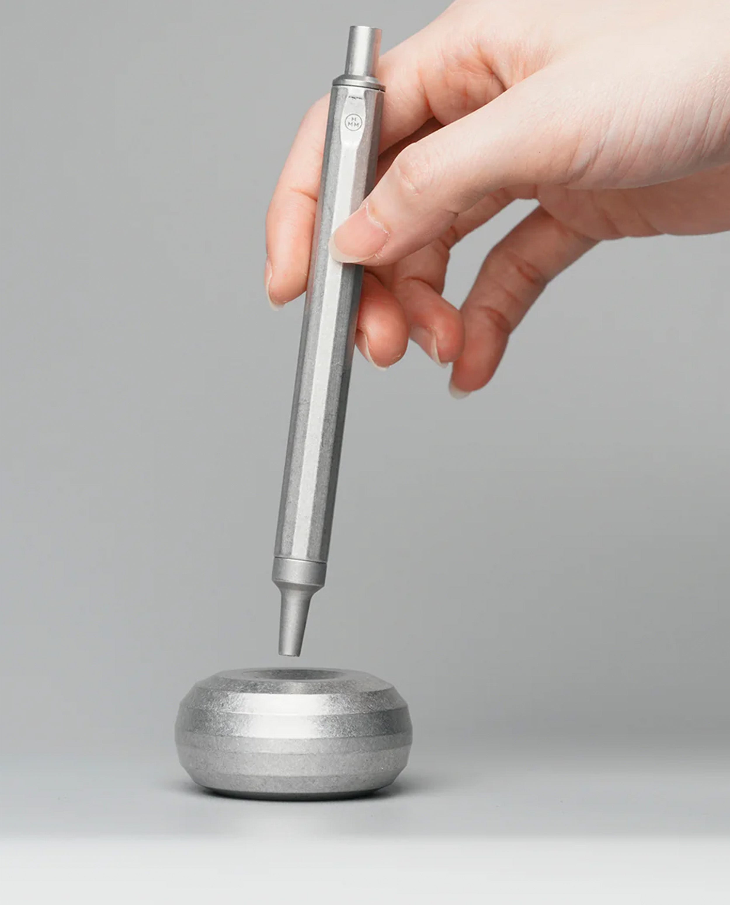 A hand holds the HMM Ballpoint Pen (Raw) vertically above a round metallic stand on a gray background.