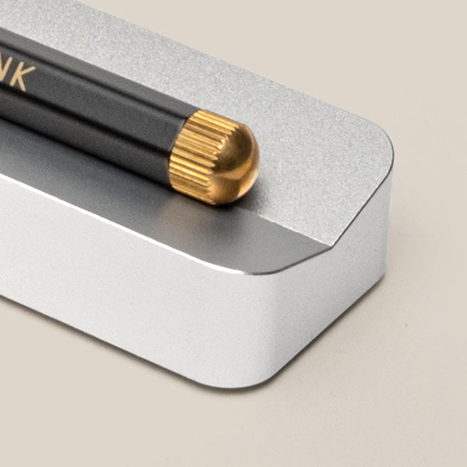 A close-up of a black pencil with a gold-colored rounded end resting on a rectangular silver metal holder, against a beige background.