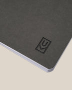 A close-up of the Ugmonk Pocket Notebooks - Set of 3, featuring a dark gray, textured soft-touch cover with a debossed logo in the lower right corner, photographed on a light beige background.