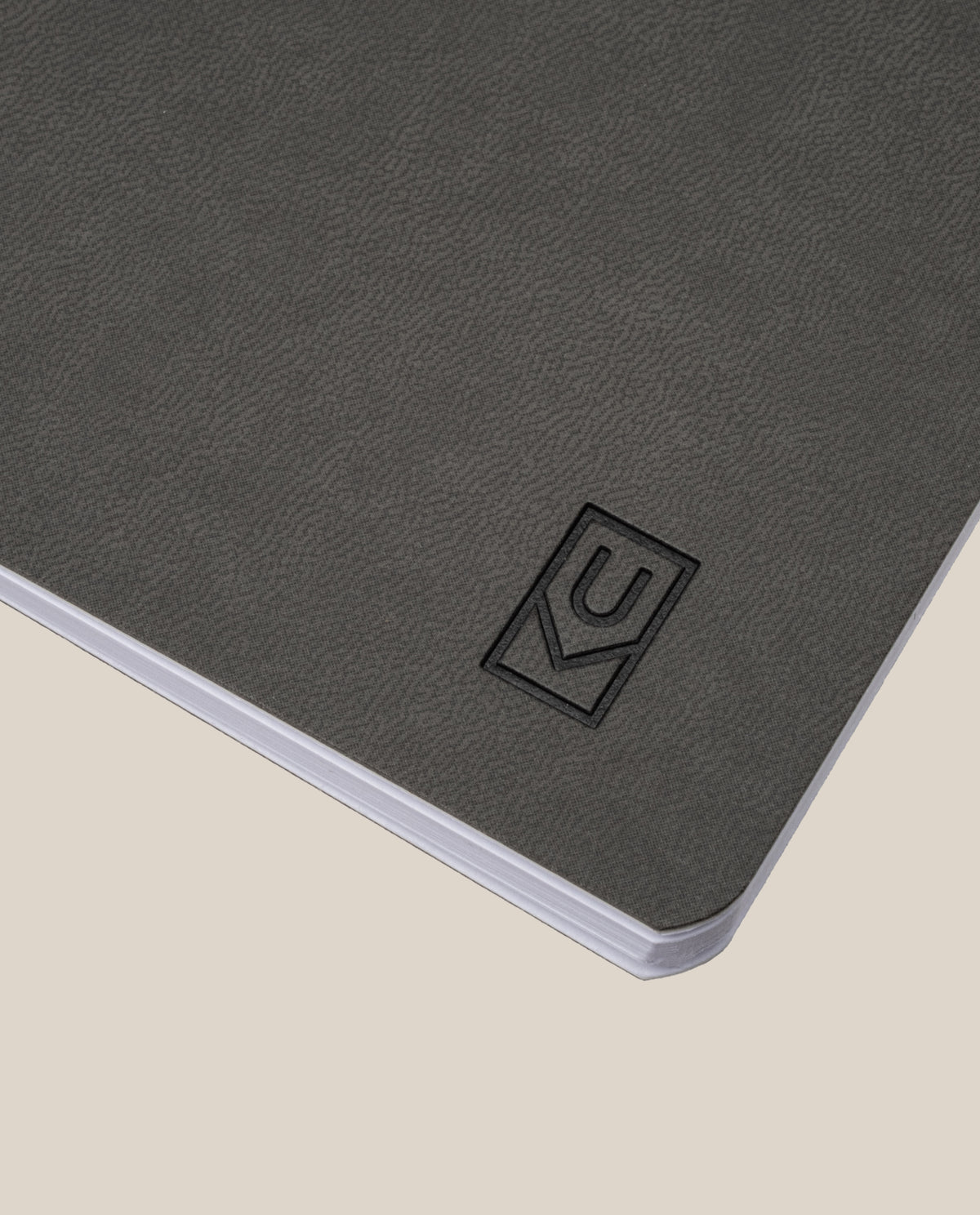 A close-up of the Ugmonk Pocket Notebooks - Set of 3, featuring a dark gray, textured soft-touch cover with a debossed logo in the lower right corner, photographed on a light beige background.