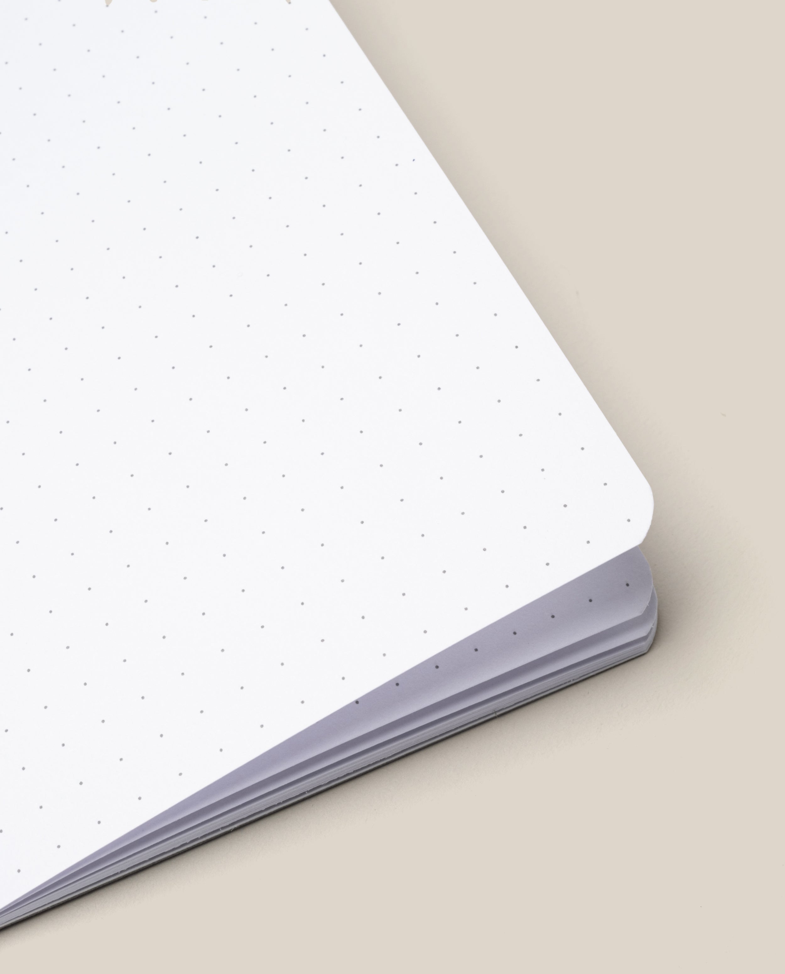 A Ugmonk Pocket Notebook from the Set of 3 rests open on a beige surface, revealing the corner of its dot grid pages.