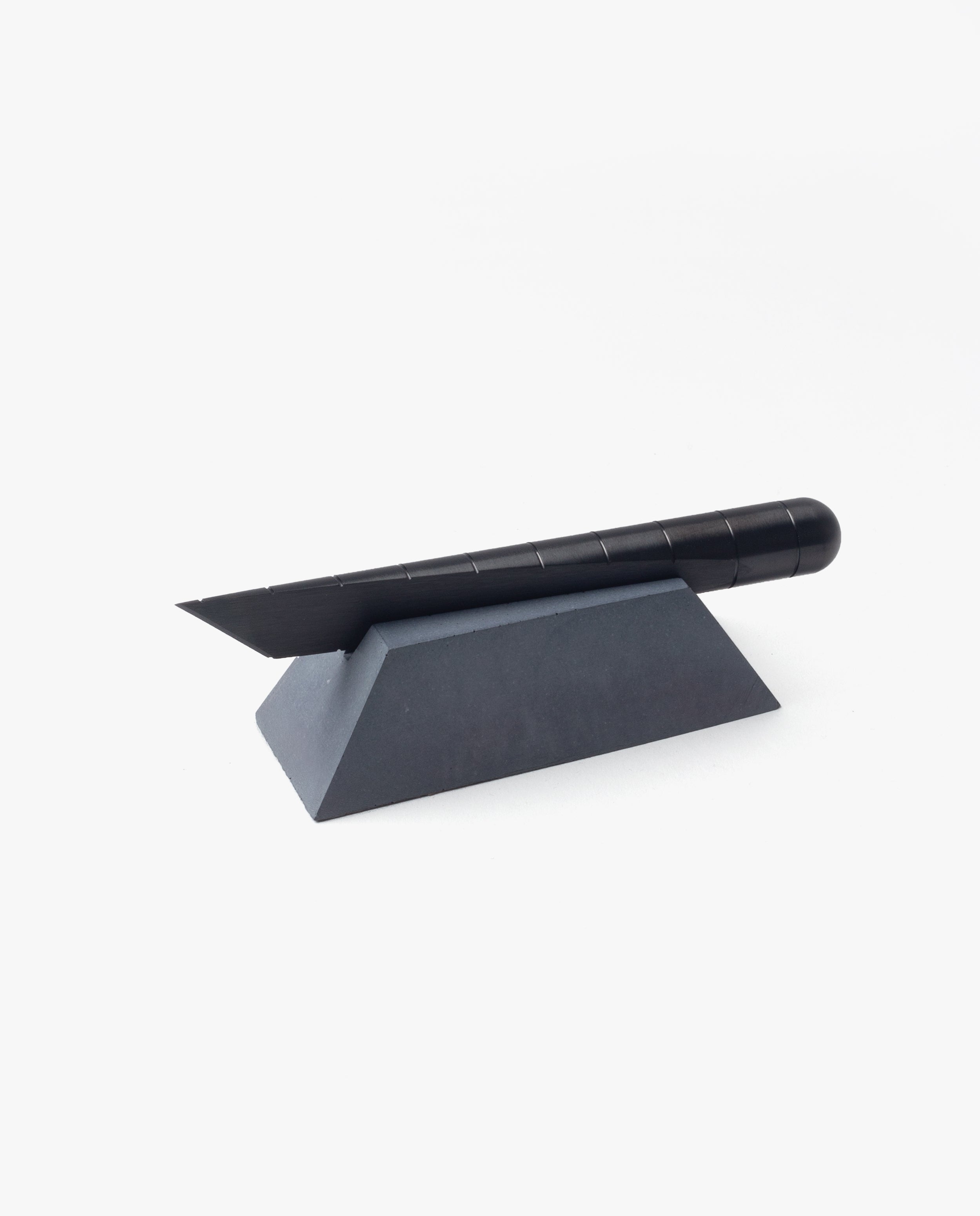 A black utility knife with a segmented blade rests on the Craighill Desk Knife Plinth (Slate), a geometric, wedge-shaped cast concrete holder, against a white background.