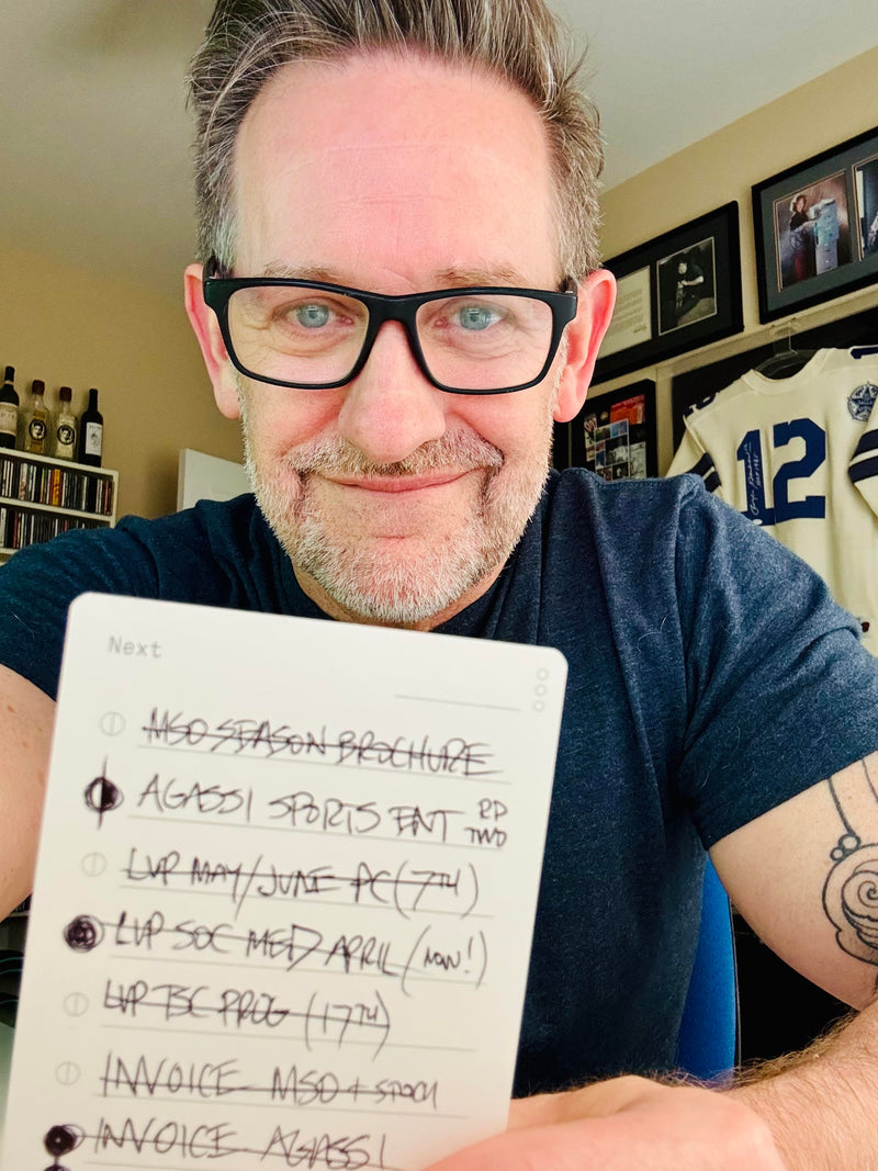 A man with glasses and a beard smiles, holding a notepad with a handwritten to-do list. Framed sports memorabilia and bottles are visible in the background.