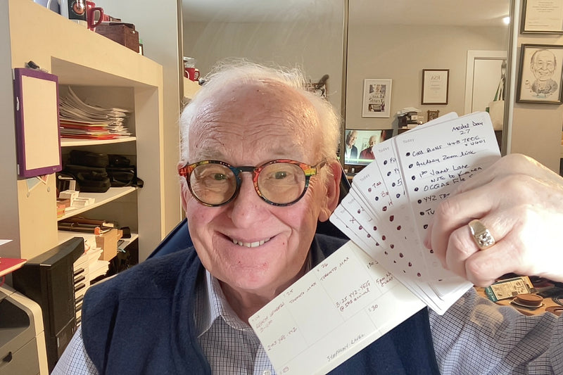 Smiling elderly man with glasses holds up several handwritten index cards in an office with shelves, papers, and a mirror reflecting the room behind him.
