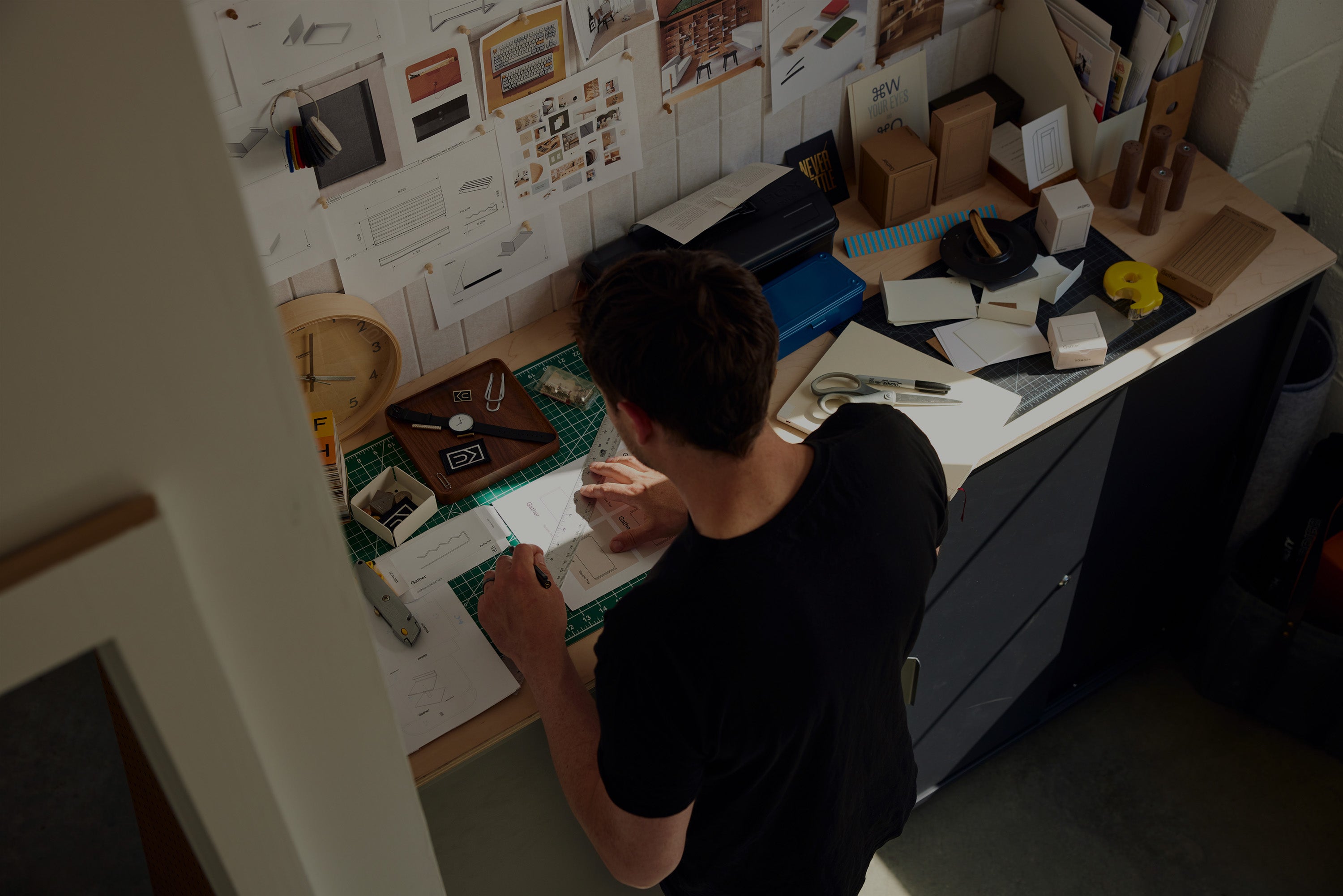 A person stands at a cluttered desk, sketching on paper. The workspace is filled with design tools, models, and materials, and the wall is covered with notes, plans, and inspiration images.