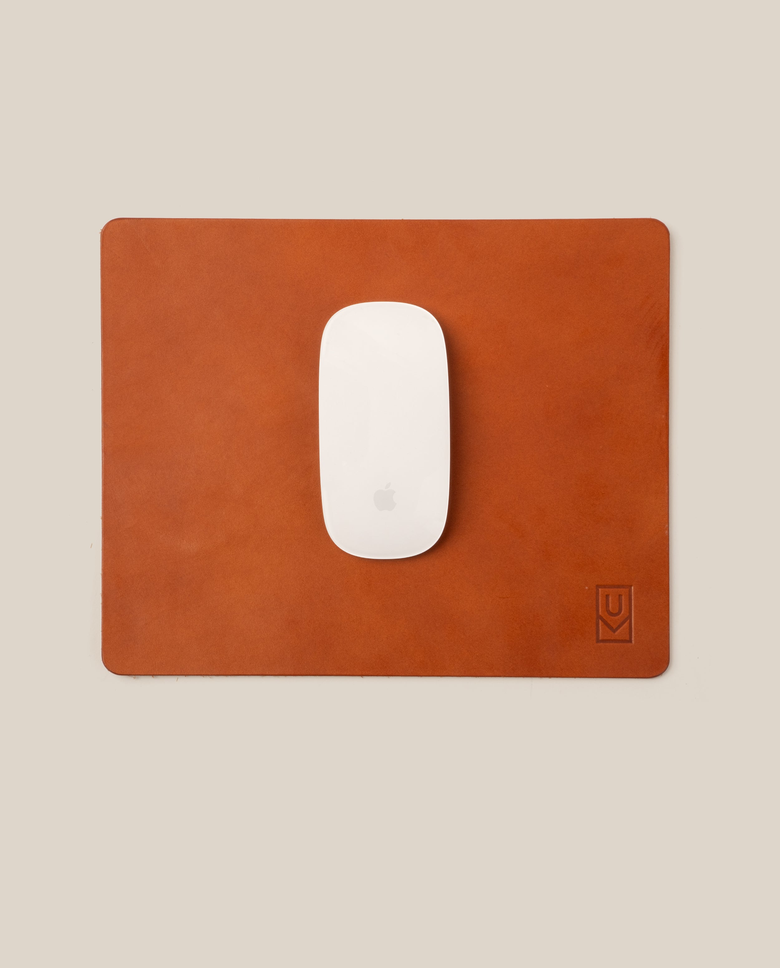 A white Apple Magic Mouse sits centered on the Ugmonk Leather Mousepad (Tan), handcrafted from full-grain leather with a subtle debossed logo in the lower right, all set against a neutral beige background.