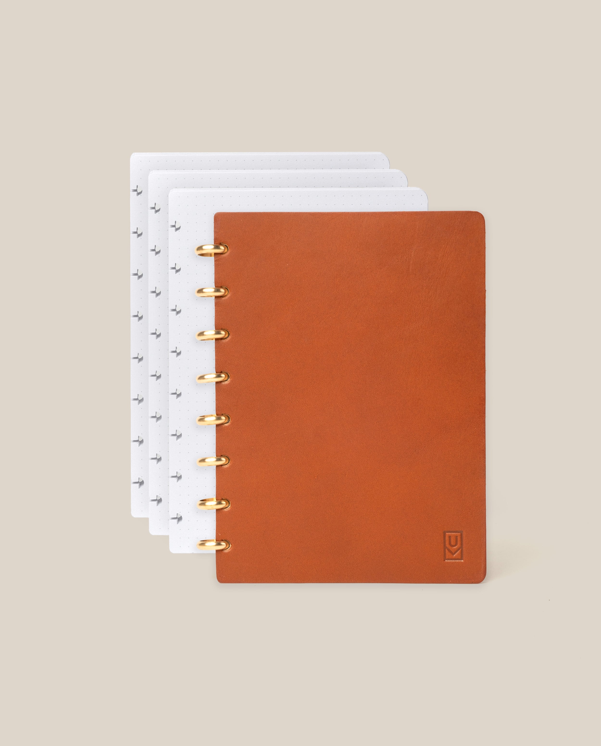 A Discbound Heirloom Journal Bundle in tan full-grain leather (standard 6" x 8.5") with gold rings, partially open to reveal dotted white pages and three refills inside, displayed on a beige background.