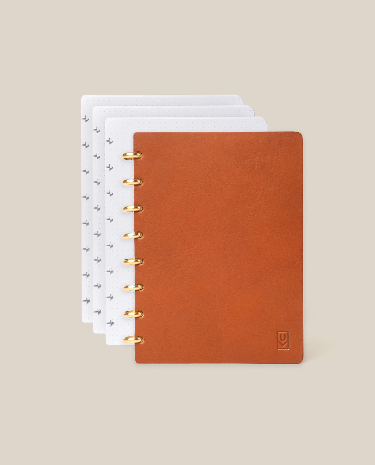 A Discbound Heirloom Journal Bundle in tan full-grain leather (standard 6" x 8.5") with gold rings, partially open to reveal dotted white pages and three refills inside, displayed on a beige background.