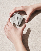Two hands positioned around a geometric, silver Craighill Tetra Puzzle (Stainless Steel) on a textured, light-colored surface, casting soft shadows.