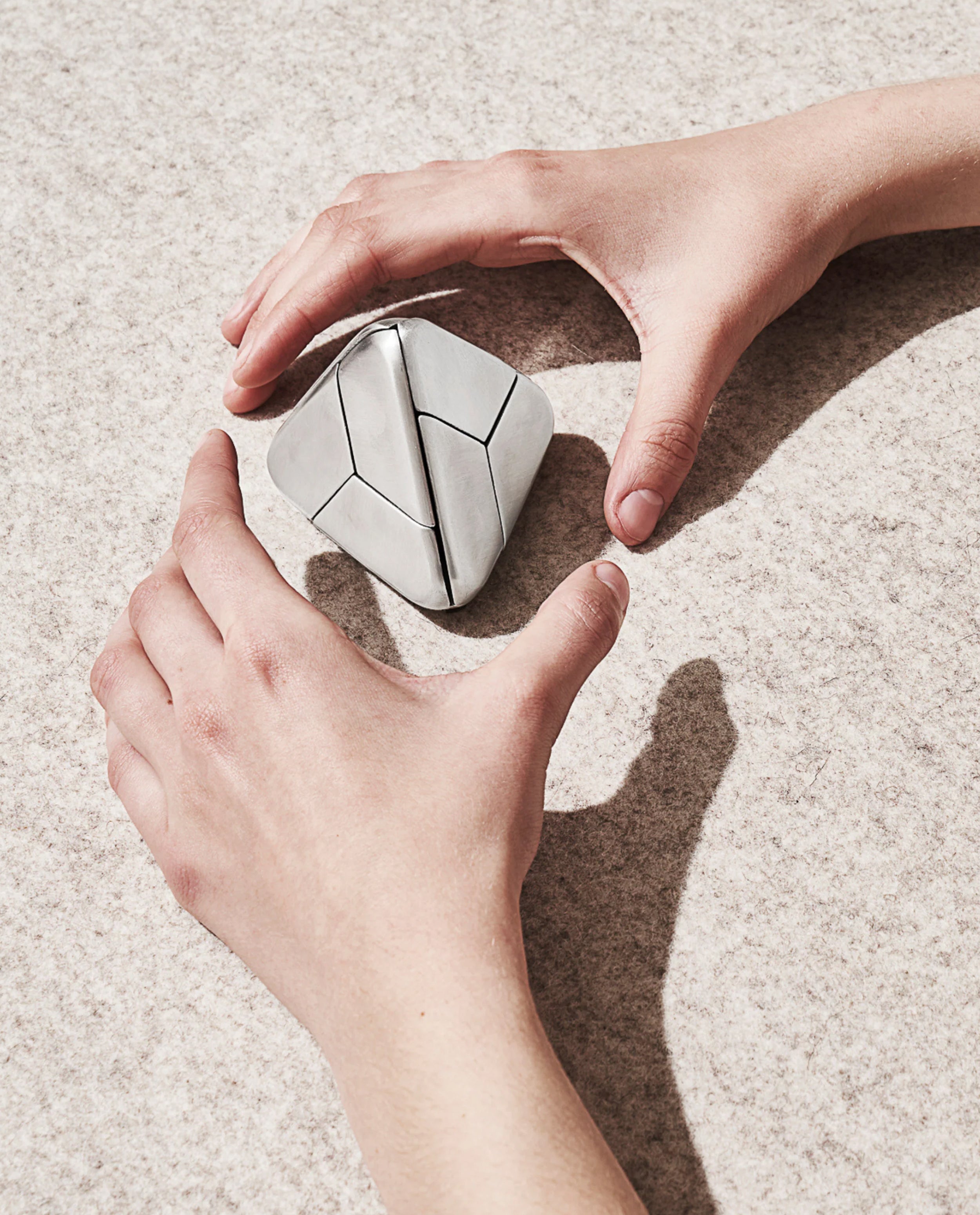 Two hands positioned around a geometric, silver Craighill Tetra Puzzle (Stainless Steel) on a textured, light-colored surface, casting soft shadows.