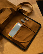 Ugmonk Utility Tote (Field Tan) canvas bag on a wooden surface with a front pocket containing two pens and a small notebook.