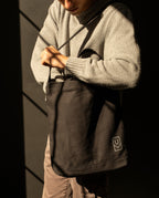A person wearing a gray sweater holds a sturdy, black Ugmonk Utility Tote (Black) with a small logo on the lower corner.