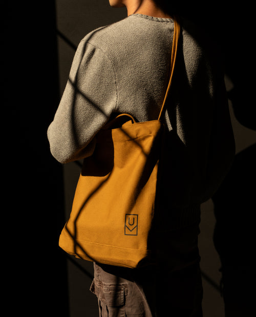 Person wearing a field tan Ugmonk Utility Tote over their shoulder, featuring a sturdy canvas construction and a square logo patch.