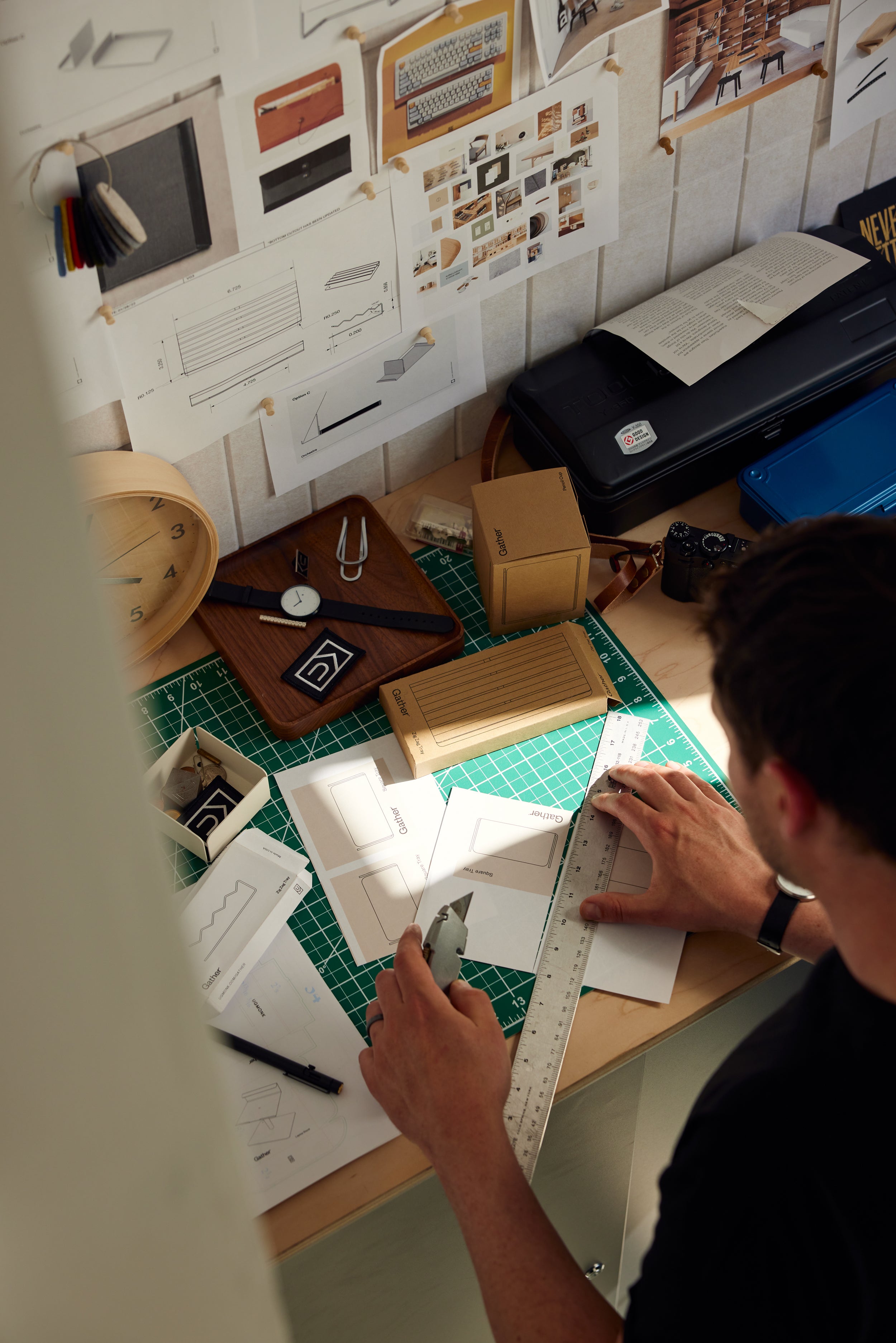 A person uses a ruler and utility knife to cut paper on a cluttered desk with design sketches, a clock, boxes, and drawing tools, in a workspace lit by natural light.