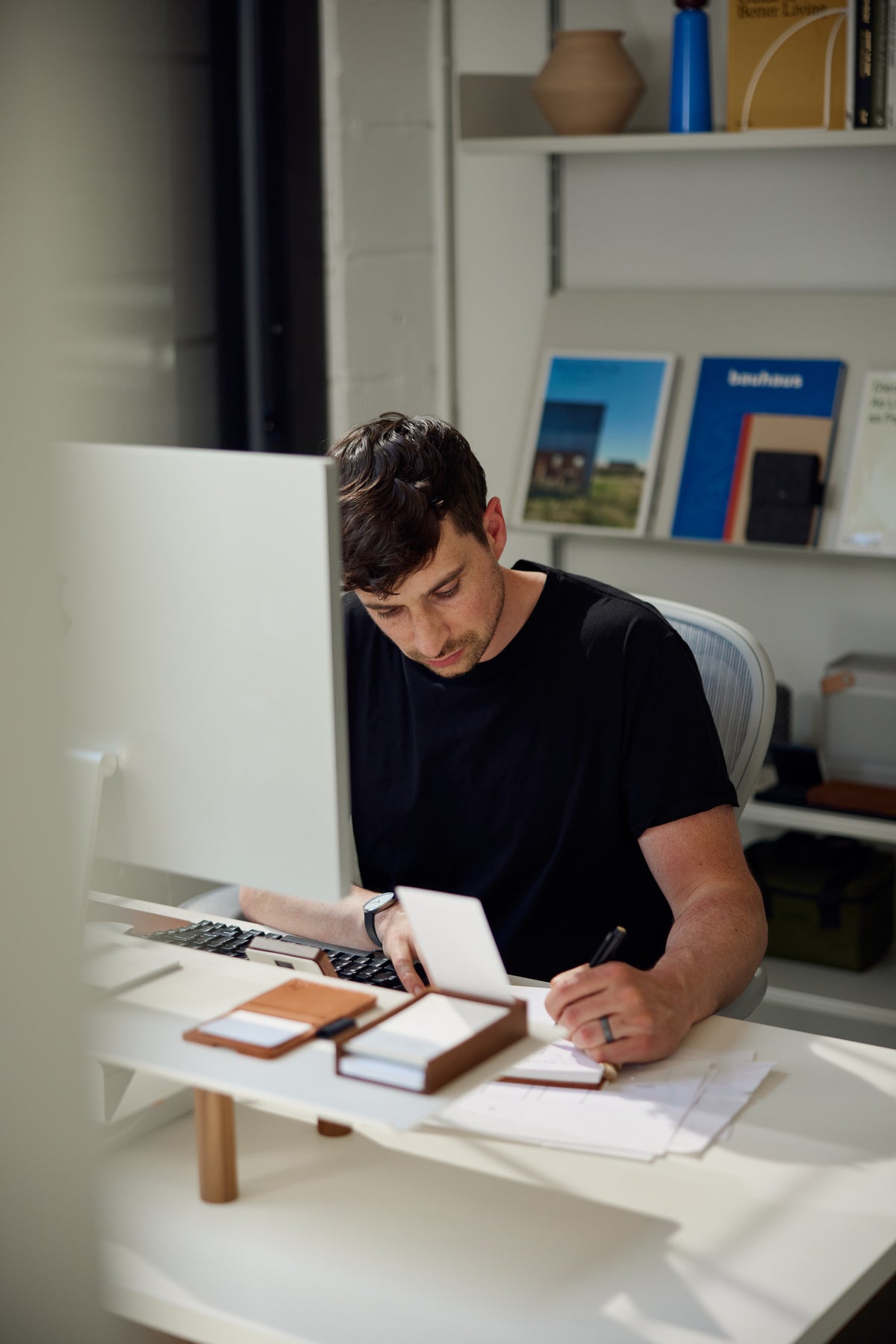 A man in a black shirt sits at a desk writing on paper, with a computer monitor in front of him and books and magazines on shelves in the background.