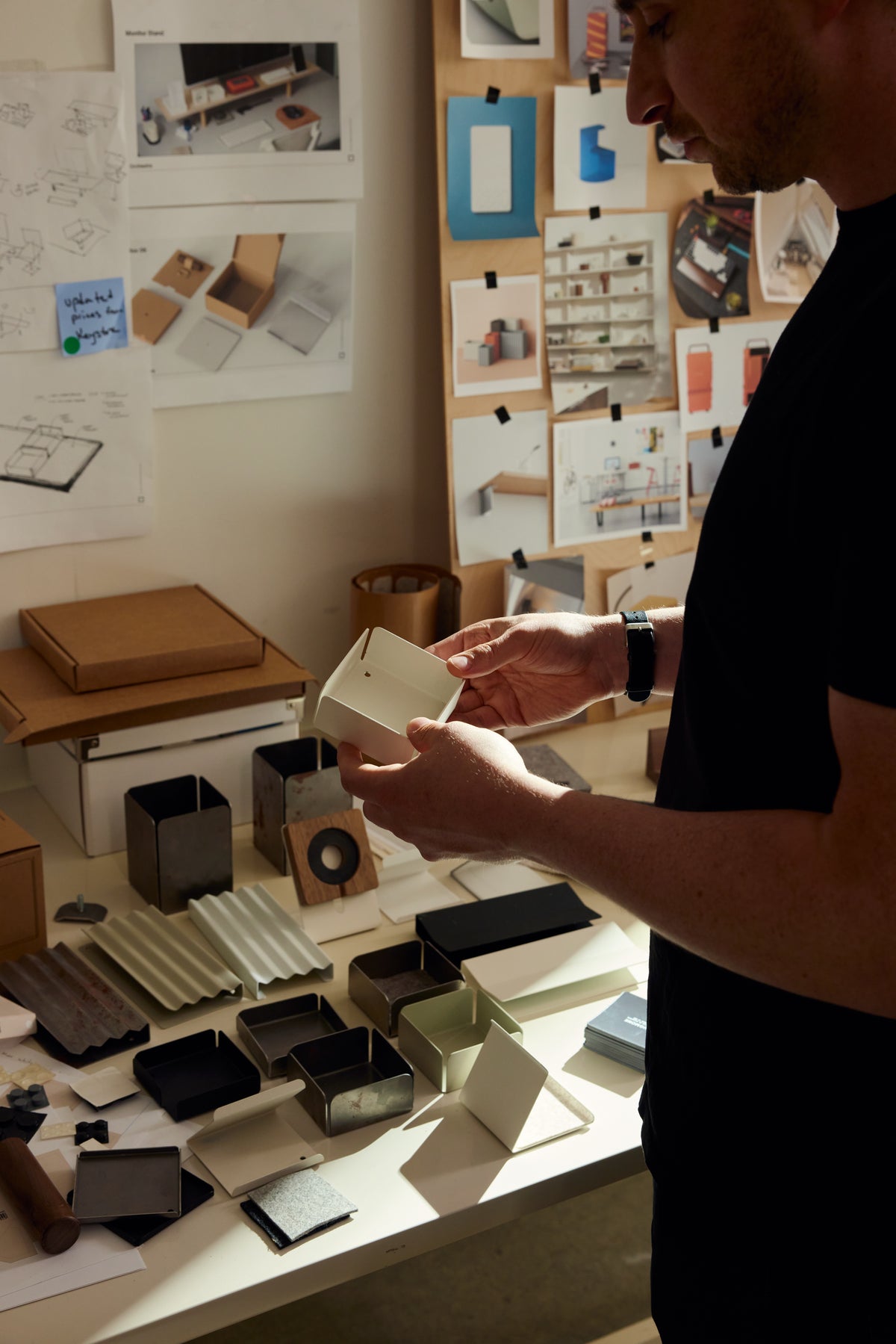 A person stands at a desk examining a small box, surrounded by various packaging prototypes, materials, and design sketches pinned to the wall.