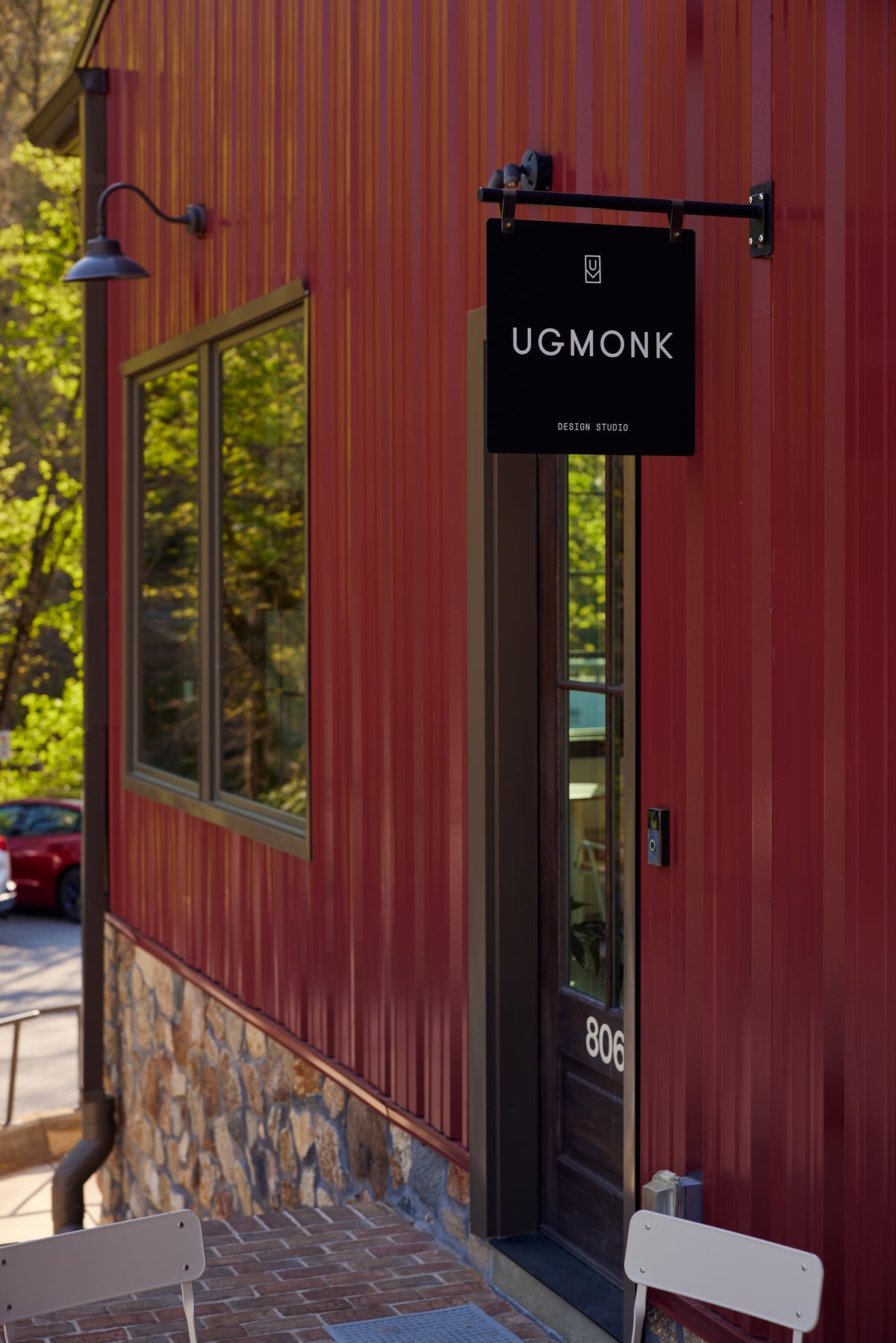 A red building with a stone base has a window and a door labeled 806. A black sign above the door reads UGMONK DESIGN STUDIO. Outdoor chairs are visible in the foreground.