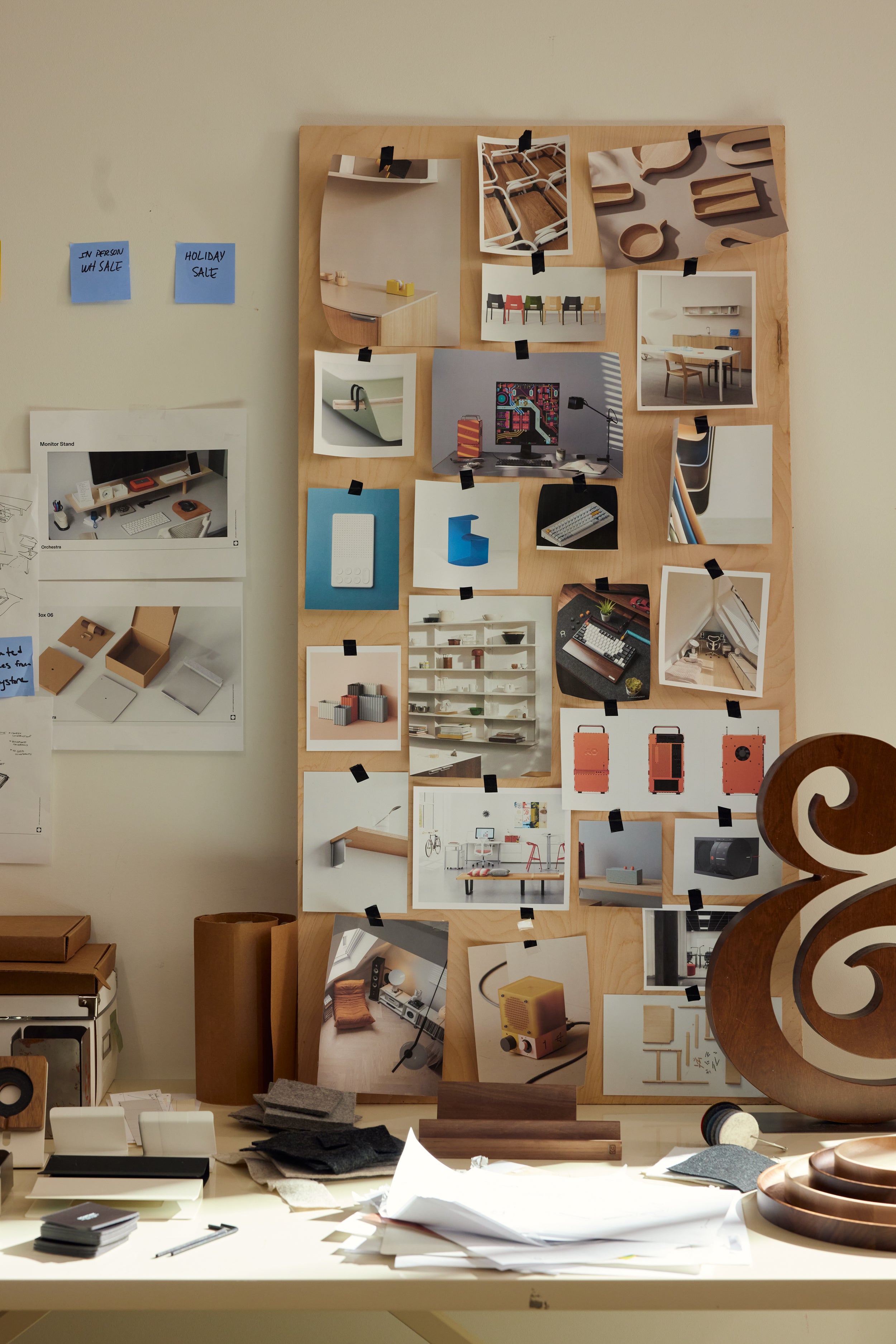 A mood board with various product and interior photos is propped on a cluttered desk with papers, samples, and a wooden ampersand decoration. Blue sticky notes and sketches are pinned to the wall beside it.