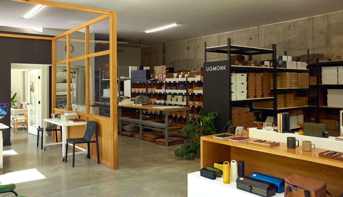 Modern workspace with shelves full of organized boxes, a wooden partition, desks with chairs, various office supplies, and plants. There is a sign that reads UGMONK near the shelves. The space has a clean, minimal design.