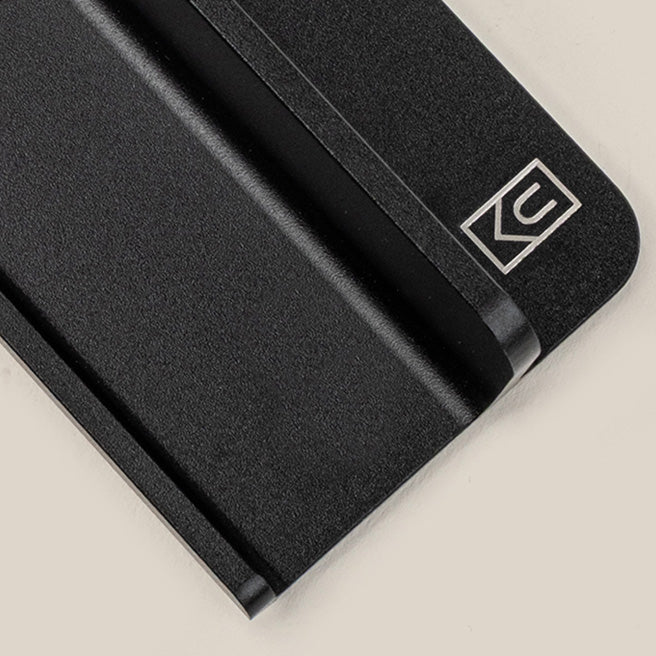A close-up of a black rectangular object with a groove, featuring a white geometric logo in the corner, on a light beige background.