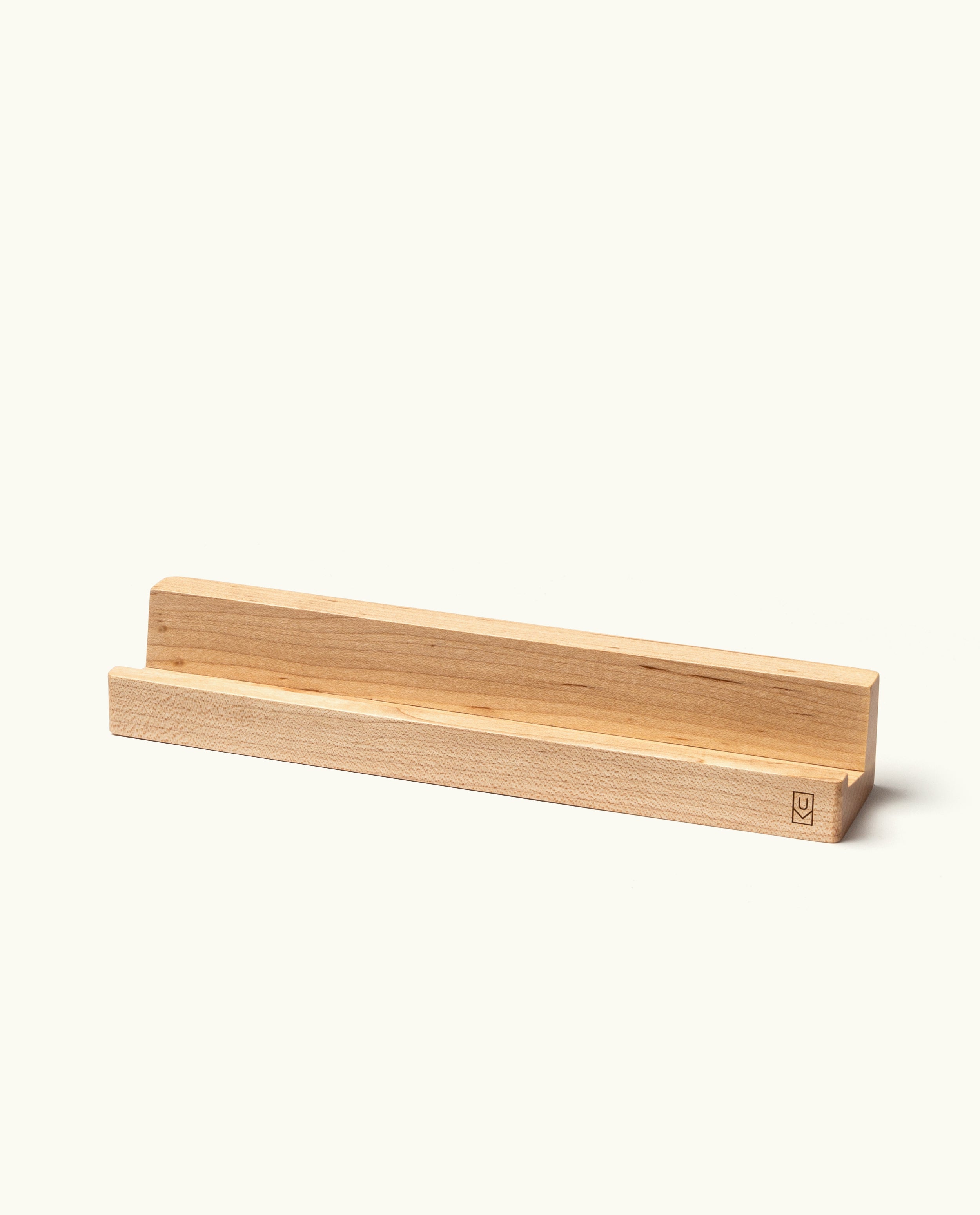 The Analog Weekly Card Holder (Maple) is a simple, rectangular stand made from light maple wood with a groove designed to hold items upright—perfect for displaying your Analog Weekly Cards. Handcrafted in Pennsylvania, this card holder features a small logo engraved on one corner. The background is plain and off-white.