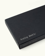 A black rectangular box with "Analog Weekly" printed in white on the lid, and "ugmonk.live/analog" on the side. Designed to house the Analog Weekly Planning Kit (Walnut) and its walnut card holder, the box is set against a plain, light background.