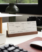 A handwritten weekly planner on a wooden stand sits on a desk, with a lamp, camera, and keyboard nearby. The desk is by a window with a blurred green view outside.