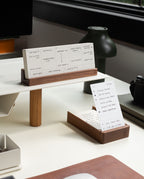 A tidy and organized modern desk featuring the Analog Weekly Planning Kit (Walnut), with its walnut card holder displaying handwritten notes and weekly planner cards. The setup is complemented by a green lamp, a black camera, and a window in the background, creating an ideal workspace for productivity.