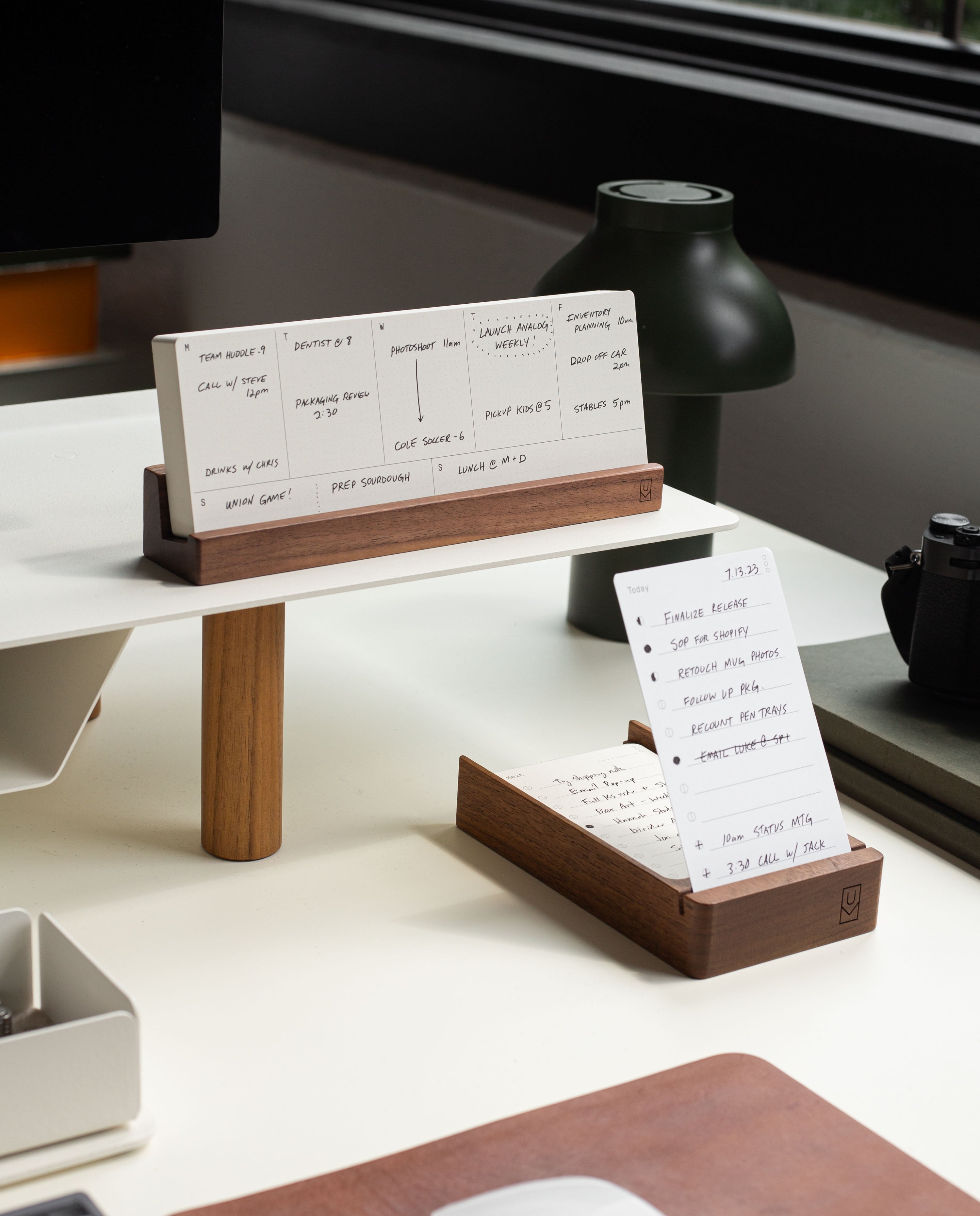 A tidy and organized modern desk featuring the Analog Weekly Planning Kit (Walnut), with its walnut card holder displaying handwritten notes and weekly planner cards. The setup is complemented by a green lamp, a black camera, and a window in the background, creating an ideal workspace for productivity.