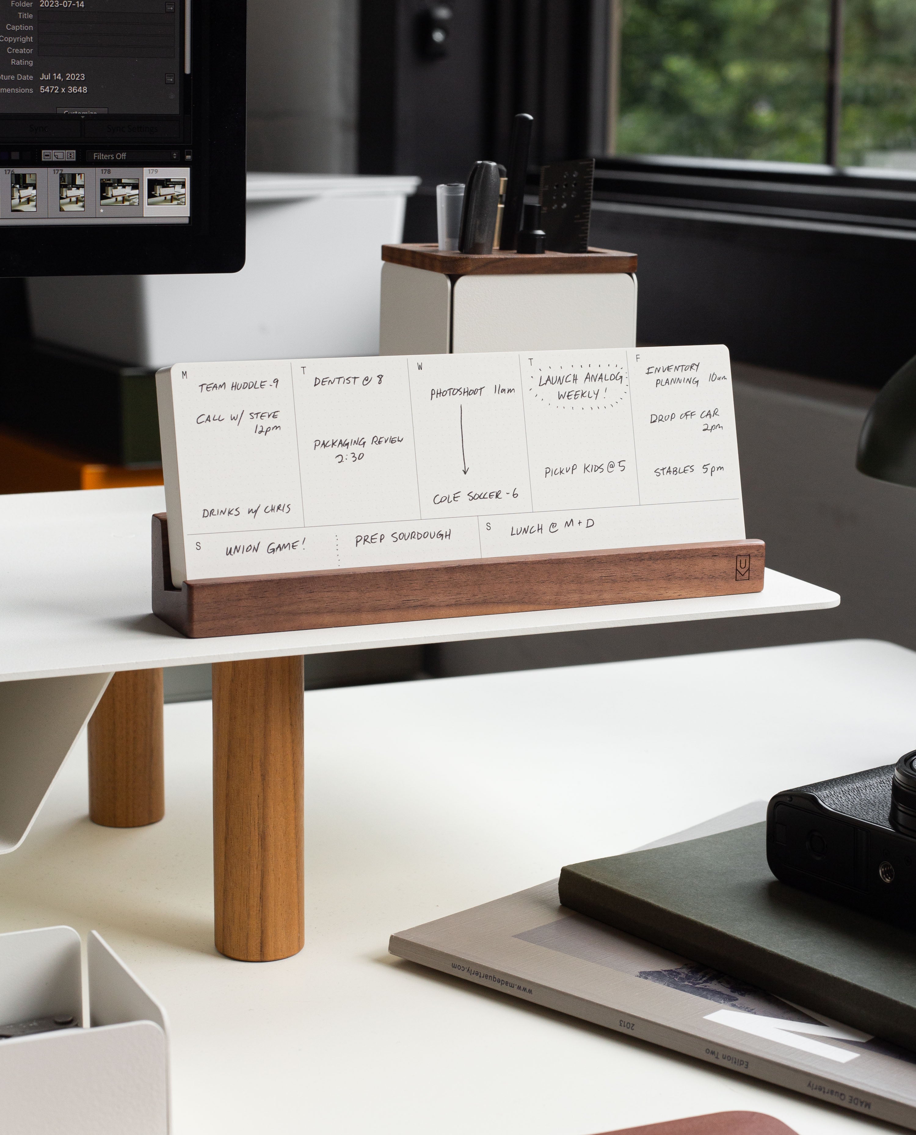A modern desk setup features a wooden organizer neatly holding handwritten white cards, pens, and a notebook. The Analog Weekly Planning Kit (Walnut) takes center stage with its sleek walnut card holder displaying weekly planner cards. In the background, a monitor, office supplies, and a window with views of greenery complete the scene.