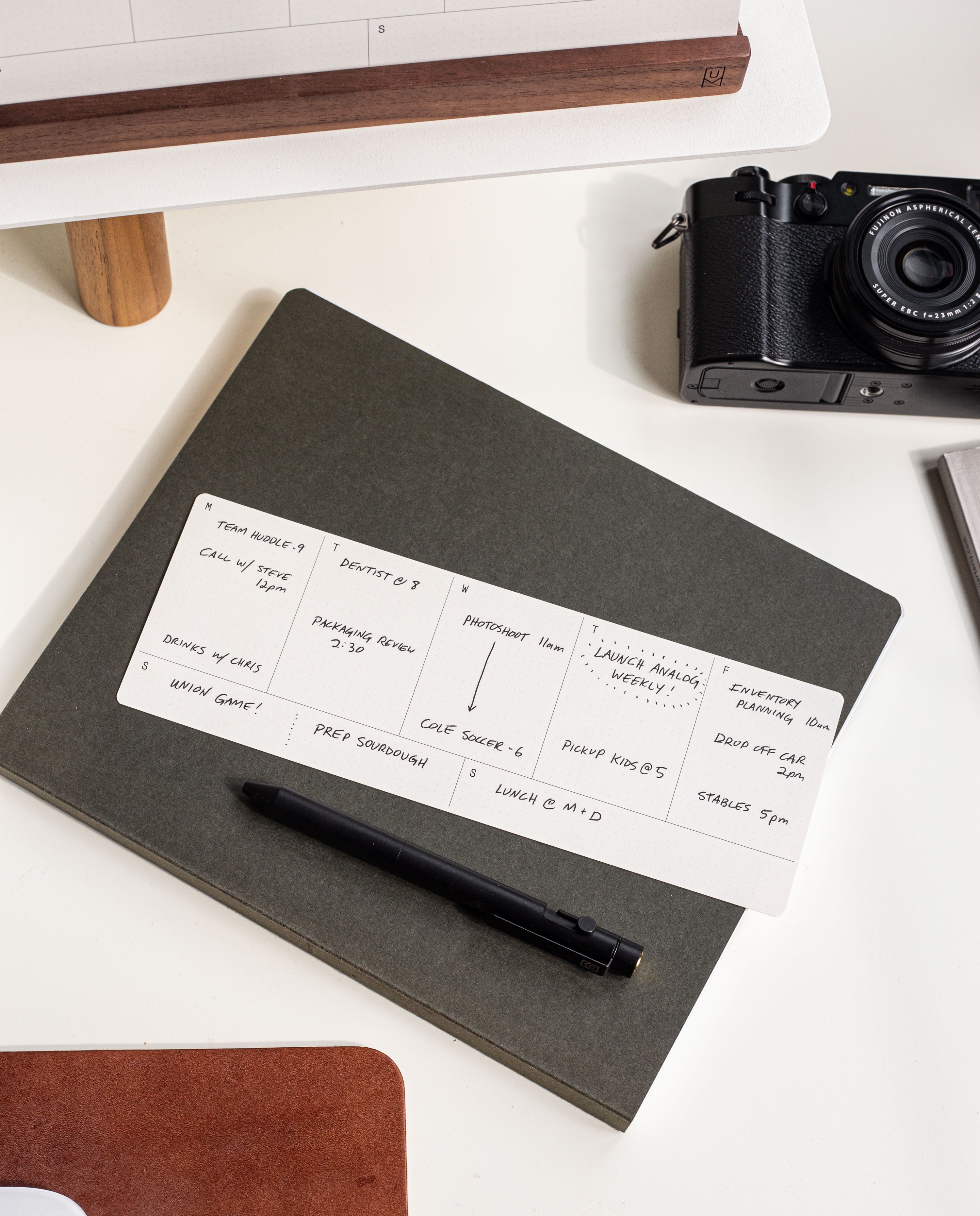 A black pen rests on a closed dark notebook, topped with a handwritten daily schedule. Nearby are a black camera, a white monitor riser, and part of a white desk. A walnut card holder elegantly displays neatly arranged weekly planner cards from the Analog Weekly Planning Kit (Walnut), adding an organized and refined touch to the workspace.
