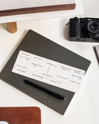 An Analog Weekly Card sits atop a closed dark notebook with a black pen, offering organized planning. In the background, a camera and part of a computer monitor are visible on the white desk.